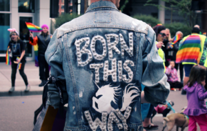 man in jean jacket with "born this way" on the back. People with rainbow flags in background.